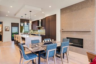 A modern open-concept dining room and kitchen with a dark marble dining table, blue upholstered chairs, and a fireplace set in a textured stone wall.