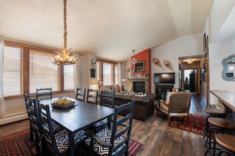 A wide shot of The Zephyr Mountain Lodge 2701's open-concept living area, featuring a dining table with an antler chandelier, a living room with a stone fireplace, and a bar area.