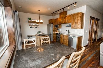 A full view of the rustic modern kitchen and dining area in Cana Loft 302, featuring wooden cabinets, stainless steel appliances, a dark granite dining table, and hardwood floors.
