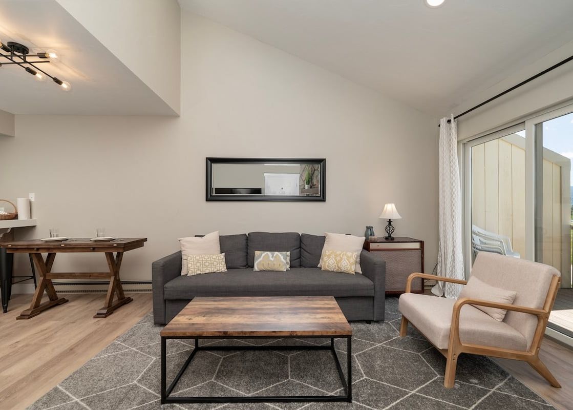 A cozy living room with a gray sofa, wooden coffee table, and light-colored armchair. A large mirror hangs above the sofa, and a glass door leads to a balcony with a mountain view.