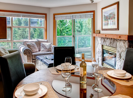 The Aspens on Blackcomb Unit 547 features a spacious living area and dining space, showcasing a large round wooden dining table set for guests with white dinnerware and wine glasses. The comfortable plaid sofa and armchair invite relaxation by the stone f