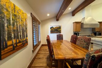 The dining area in The Chateau Deluxe Four Bedroom Suite features a long wooden table with patterned chairs, a large painting of golden trees on one wall, and a kitchen area visible in the background.