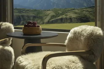 Two plush, white sheepskin chairs with wooden frames flank a small black table, on which a wooden bowl of red apples rests, all set against a large window revealing a panoramic view of green hills and towering mountains under a clear blue sky.