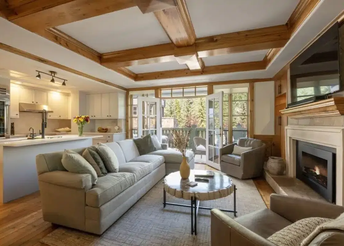 A luxurious open-concept living area at Chateau Beaver Creek Residence 202 featuring a comfortable sofa, a gas fireplace, and a kitchen with an island, all brightly lit by natural light from large French doors leading to a balcony.