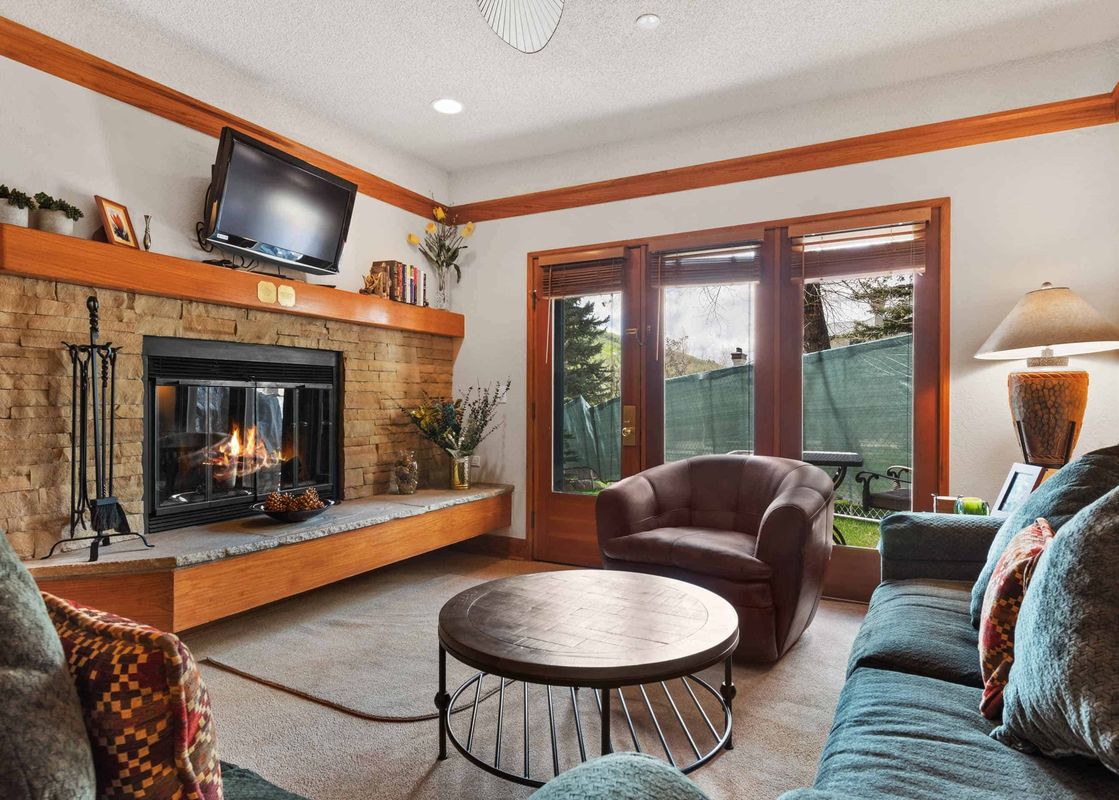 A cozy living room featuring a stone fireplace with a TV mounted above, a dark green sofa, a brown armchair, and a circular coffee table, with large windows overlooking an outdoor scene.