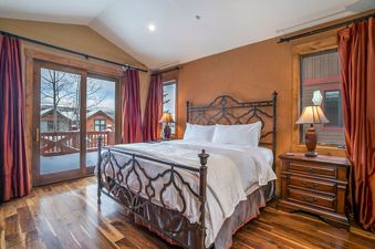 The bedroom at Hygge Hideaway boasts a large bed with a decorative metal frame, white bedding, and two nightstands with lamps, all set on a polished hardwood floor with a view of snow-covered trees outside the sliding glass door.