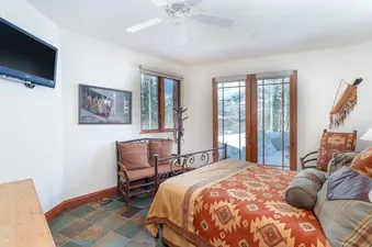 A cozy bedroom at Pine Meadows 138 features a bed with a patterned duvet, a wall-mounted TV, tile flooring, and French doors leading to a snowy mountain view.
