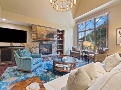 A cozy and elegant living room with a stone fireplace, large window overlooking a forest, comfortable seating, and a vibrant blue rug.