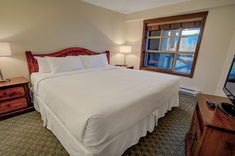 A cozy bedroom in Blackcomb Springs One Bedroom Slopeside features a comfortable king-size bed, two nightstands with lamps, a window, and a TV on a dresser.