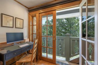 A home office with a black desk, a monitor, and a wooden chair, with French doors opening to a balcony with green railings and a view of green trees.
