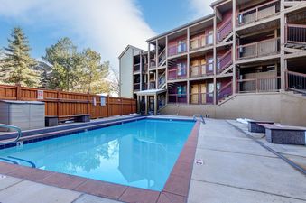 An inviting outdoor pool with clear blue water is set against a multi-story building at Snowcrest 205, surrounded by a wooden fence and lush evergreen trees under a bright sky.