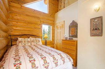 The bedroom at Miles Behind features a cozy bed with a floral duvet, wooden walls, a dresser, and two windows offering views of the outdoors.