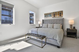 A bright and modern bedroom at Apex 1501 features a grey upholstered bed with patterned bedding, two nightstands, and two windows looking out onto city buildings.