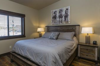 A king-sized bed with gray linens is flanked by two lamps, with a horse painting above the headboard and a window to the left.