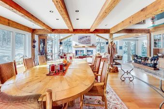 The Trails Edge Vacation Home's dining area features a large wooden table with chairs, set in front of windows overlooking a snowy landscape, with a living room and fireplace visible in the background.