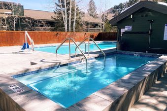 An outdoor hot tub and pool area, featuring sparkling blue water, surrounded by a stone patio and lush trees under a clear sky.