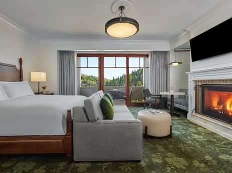 The Montage Deer Valley King Room Vista View features a king-sized bed with white linens, a comfortable seating area, a fireplace, and a private balcony overlooking a lush, forested mountain landscape.