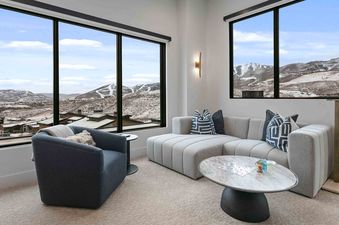 A cozy living area with large windows showcasing snow-covered mountains, featuring a light gray sectional sofa, a marble coffee table, and a blue armchair.