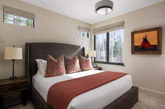 A king-sized bed with a rust-colored throw and patterned pillows dominates a bedroom at Moonlight Basin Lodging 7 Rolling Ridge, featuring large windows with forest views and a painting of a teepee.