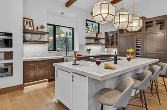 A modern and luxurious kitchen with dark wood cabinets, white marble countertops, and a large island with bar stools, accented by contemporary lighting and an exterior view.
