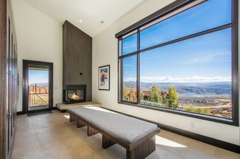 A spacious room features a modern fireplace, a long bench, and a large window offering panoramic views of a lake and mountains.