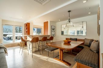 The dining area and bar of Le Chamois Penthouse, featuring a wooden table with bench seating, a breakfast bar with stools, and large windows looking out to a snowy landscape.
