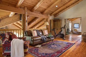 A rustic living room with log beams on the ceiling, featuring a brown leather sofa, a patterned armchair, a Peloton bike, and a colorful area rug, with a stone fireplace visible in an adjacent room.