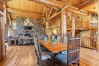 A rustic dining room with a large wooden table and chairs, a fireplace in the background and a loft area above.