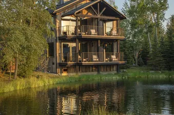 A multi-story wooden lodge with balconies overlooking a calm lake, surrounded by green trees and grass under a clear sky.