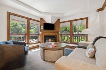 A cozy living room in The Aspen 44ABC with a lit fireplace, comfortable seating, and large windows showcasing a green outdoor view.