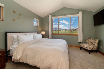 A cozy bedroom features a bed with white linens, a bedside table with a lamp, a patterned armchair, and large windows offering a view of mountains and a field.