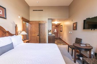 A bedroom with a king-size bed, a small table, a flat-screen tv, and a door leading to a hallway.