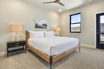 A modern bedroom with a king-sized bed, two nightstands with lamps, a window with a mountain view, and a ceiling fan.