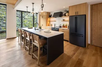 The modern kitchen in The Sylvan Lodge One Bedroom Penthouse features a large island, black appliances, and floor-to-ceiling windows overlooking a natural landscape.