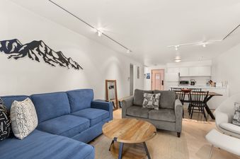 A bright open-concept living room and kitchen with a blue sectional, gray loveseat, round coffee table, and a dining area with a kitchen featuring white cabinets and appliances.