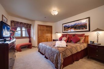 The Chateau Chamonix CX133's bedroom features a king-sized bed with a red and gold comforter, a framed mountain landscape painting, and an upholstered bench by the window.
