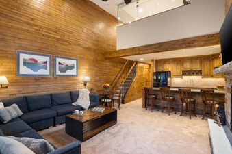 A wide view of the open-concept living room and kitchen at Montaneros 414, featuring a large sectional sofa, coffee table, breakfast bar, and staircase.