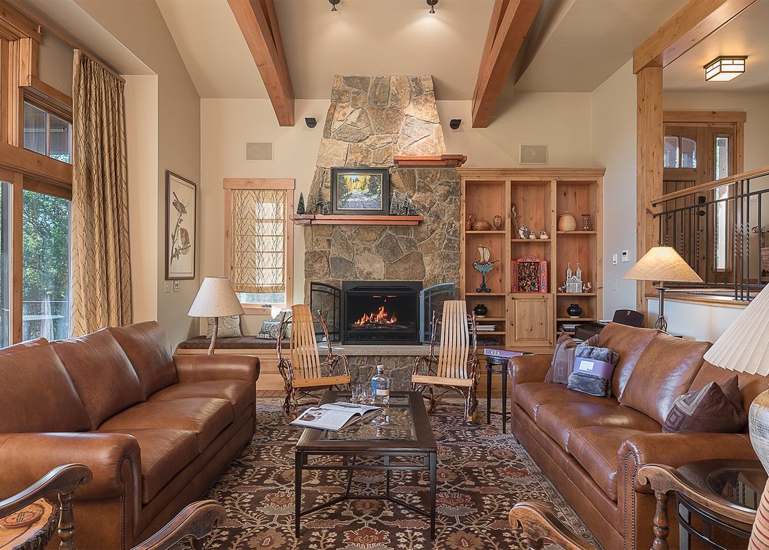 A spacious living room at Valley View Retreat with two brown leather sofas facing a large stone fireplace, showcasing rustic wooden beams, built-in bookshelves, and a view of lush trees outside.