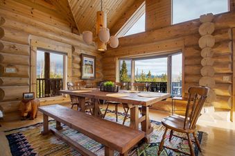 The dining area at Miles Behind features a large wooden table and bench seating, set on a vibrant patterned rug, with expansive windows offering views of the surrounding natural landscape and a warm, inviting log cabin interior.