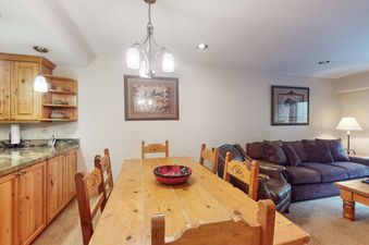 A rustic wooden dining table with six chairs sits in the dining area of Fallridge 402, with a cozy living room and a kitchen with light wood cabinetry and granite countertops visible in the background.
