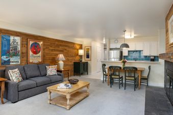 A welcoming living room and kitchen in Montaneros 311 with a gray sofa, wooden coffee table, and a dining area, featuring a wooden accent wall and a kitchen with white cabinets and a blue tiled backsplash.