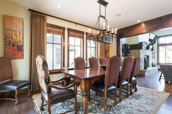 A spacious dining room features a large wooden table surrounded by eight upholstered chairs, illuminated by a rustic chandelier, with large windows and a view of a stone fireplace in the adjacent living area.