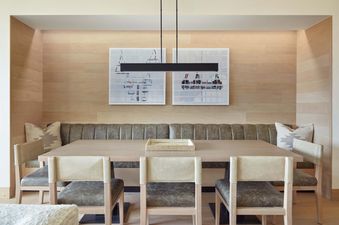 A modern dining area features a long light wood table, a dark gray upholstered banquette, light wood chairs, a sleek black linear pendant light, and two abstract artworks on a light wood paneled wall.