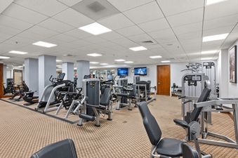 A wide shot of a modern fitness center with various workout machines, including ellipticals, stationary bikes, and weight machines, set on a light brown patterned carpet with two wall-mounted televisions.