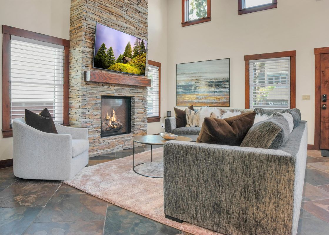 A cozy lounge at The Peak at Town Lift features a stone fireplace with a TV above, flanked by comfortable grey seating and a round coffee table on a light rug, set against a backdrop of sunlit windows.