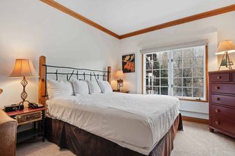 A primary bedroom in The Charter D2205 features a king-sized bed with white linens, two nightstands with lamps, a dresser, and a window with an exterior view.