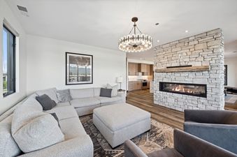 A bright and elegant living room with a large light gray sectional sofa, a stone fireplace with a linear firebox, and a stylish chandelier, offering a glimpse into an adjacent modern kitchen.
