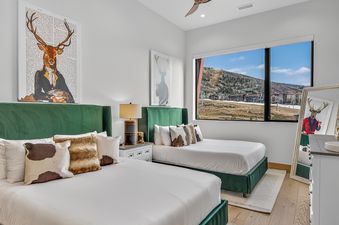 A spacious bedroom at The Duet Grande with two beds featuring green headboards, decorative cowhide pillows, a nightstand, and a large window overlooking scenic mountains.