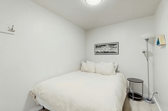 A serene bedroom featuring a bed with white linens, a mountain landscape painting, a black side table, a floor lamp, and a coat hook.