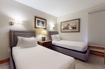 A cozy bedroom with two twin beds featuring gray upholstered headboards, a shared wooden nightstand with a lamp, and framed artwork on the walls.